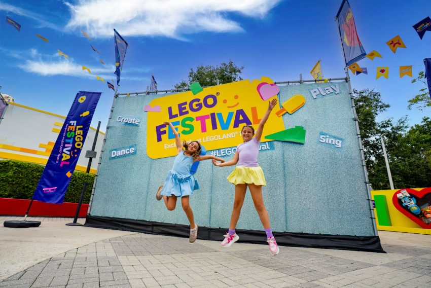 Two smiling girls in pastel dresses jump in front of a colorful LEGO Festival backdrop under a blue sky with banners and kites.",