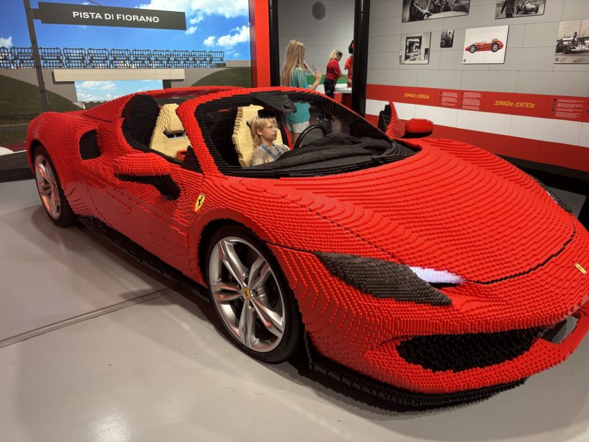 Life-size red Ferrari built from LEGO bricks on display, with silver wheels and Ferrari logo, in a showroom.
