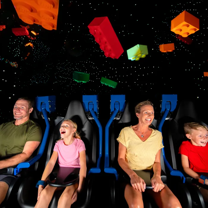 Family of four on a space-themed roller coaster, laughing in blue seats with colorful floating blocks overhead.