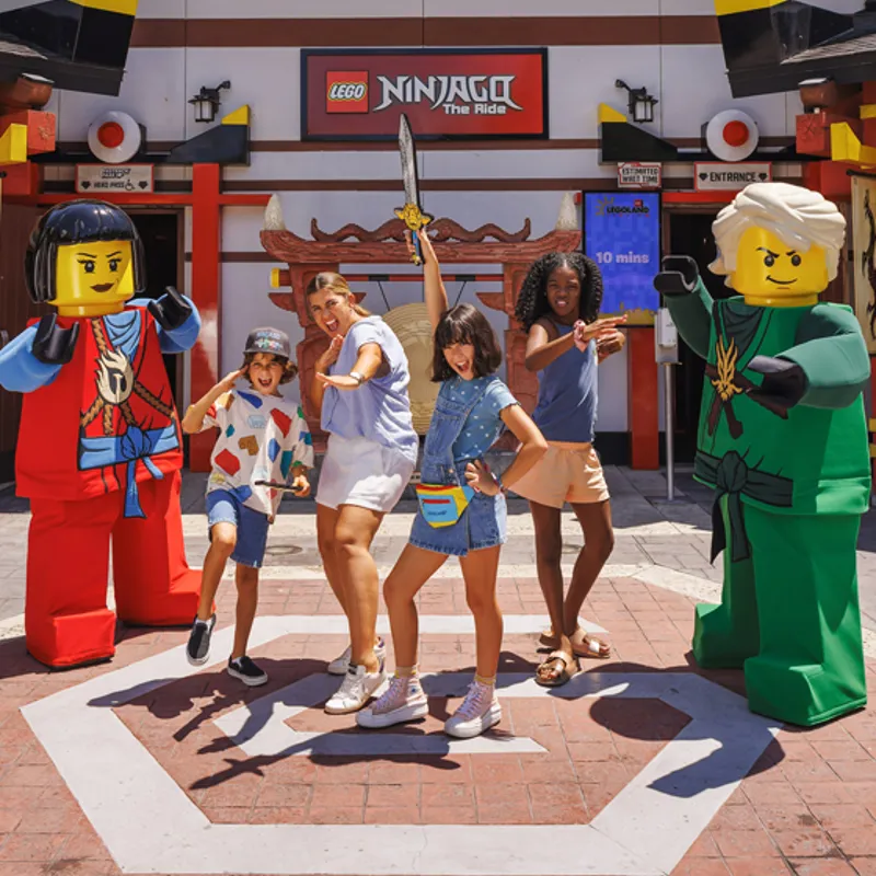 Group of kids posing with raised swords between two large LEGO minifigures outside the LEGO Ninjago The Ride entrance.